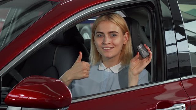 Young beautiful cheerful woman smiling to the camera while sitting in her new automobile. Happy female driver showing thumbs up holding car keys. Transportation, ownership concept.