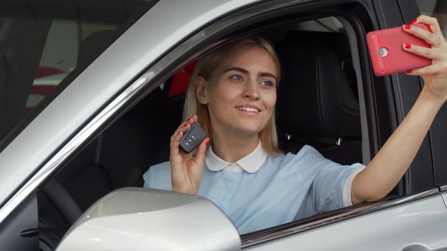 Attractive young happy woman using her smart phone while taking selfies sitting in her new automobile, holding car keys. Beautiful female driver using her phone. Technology, purchase, drive concept.