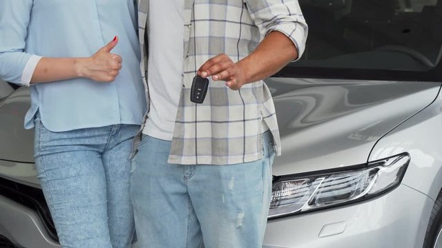 Cropped shot of a loving couple hugging, standing in front of the new automobile at the dealership. Couple holding car keys and showing thumbs up after buying a new auto. Ownership concept.
