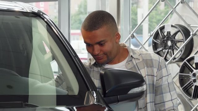 Cheerful handsome African man examining his newly bought car at the dealership showroom. Happy male driver smiling to the camera holding car keys. Buying car, driving, insurance concept.