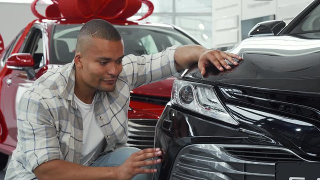 Young handsome African man smiling to the camera showing thumbs up, while examining an automobile for sale at the local dealership. Male customer choosing new auto to buy. Driving concept.