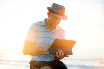 Senior man working on his laptop on the beach during sunset, freelancer concept in travel