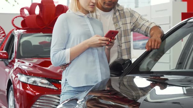 Cropped shot of a young couple using smart phone at the dealership showroom. Young people choosing new automobile to buy. Consumerism, technology, family vehicle concept.