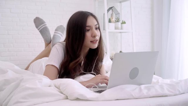 Beautiful Asian woman playing computer while lying on the bed in her bedroom. Happy female listening music with headphones and searching songs in a laptop at home. Lifestyle woman at home concept.