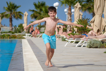 Smiling Caucasian boy having fun in swimming pool at resort on family vacation. He is running along pool edge.