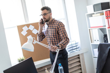 A man is standing in the office near the window and talking on the phone.