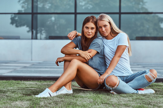 Two smiling girls cuddling while sitting on skateboard at a lawn on a background of the skyscraper.