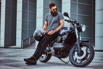 Brutal bearded male in a gray t-shirt and black pants holds a helmet sitting on his custom-made retro motorcycle against skyscraper. © Fxquadro