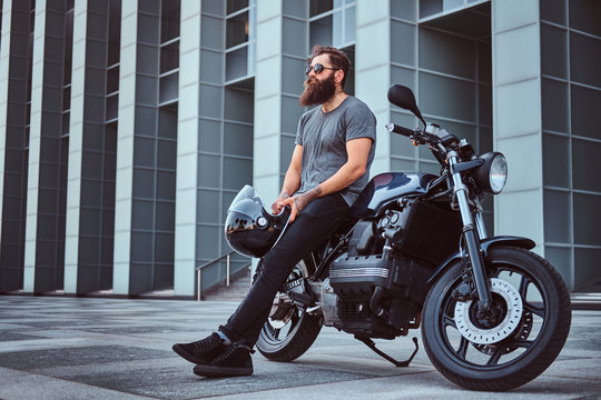 Brutal bearded male in a gray t-shirt and black pants holds a helmet sitting on his custom-made retro motorcycle against skyscraper.