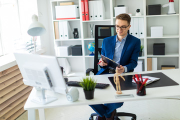 A young man sits at a table in the office and holds documents and a pen in his hands.