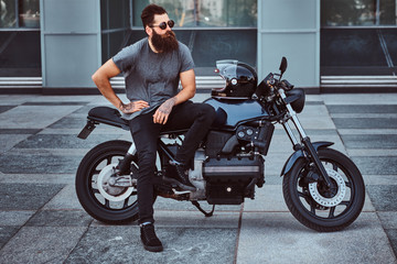 Brutal bearded male in sunglasses dressed in a gray t-shirt and black pants sitting on his...