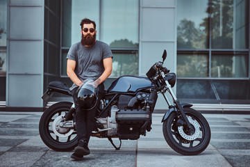Brutal bearded male in a gray t-shirt and black pants holds a helmet sitting on his custom-made...