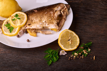Baked fish with lemon, spices and herbs on white plate on wooden background without head