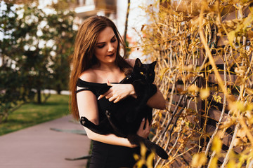charming long-haired beautiful woman in black dress holds a black cat on her hands, walks in the...