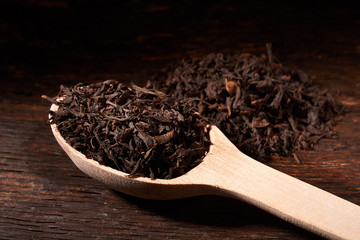 Leaves of black tea in wooden spoon on dark wooden table on black background