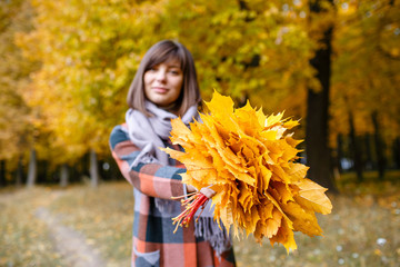 Focus on yellow leaves. Brunette woman in autumn park with fashionable plaid coat and scarf.