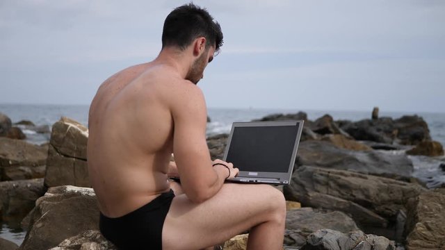 Young handsome man working on laptop computer, typing on keyboard while at the beach in front of the sea