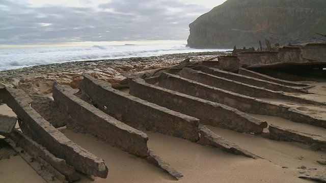 Rusty ribs as silent remains of a stranded ship's hull on a sandy beach in in front of steep cliffs and a calm surf in the afternoon sunlight