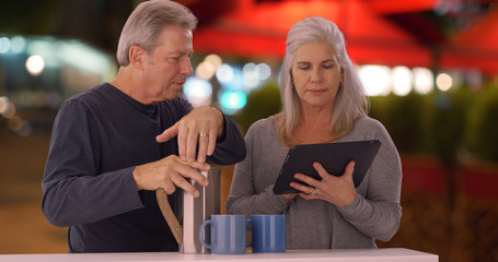 Old white couple looks up how to make coffee on tablet at outdoor cafe