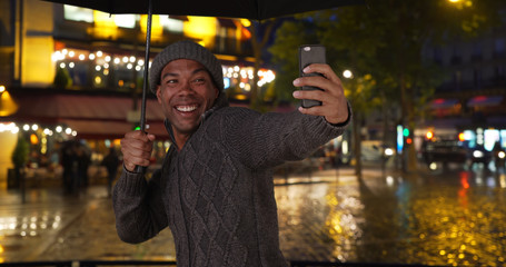 Cheerful black male taking photos of himself on rainy night out in the city