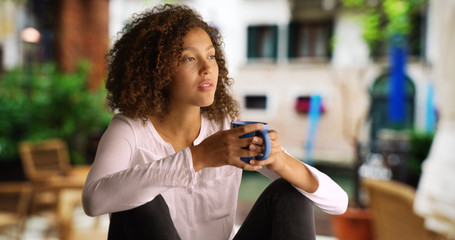 Pretty black woman with curly hair sits outside drinking coffee - Powered by Adobe