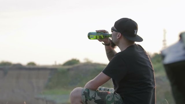 A carefree, young, friendly dude sits on a rock above the sea and drinks beer. He's wearing a black t-shirt and shorts and a black baseball cap.