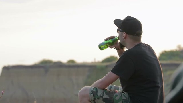 A carefree, young, friendly dude sits on a rock above the sea and drinks beer. He's wearing a black t-shirt and shorts and a black baseball cap.
