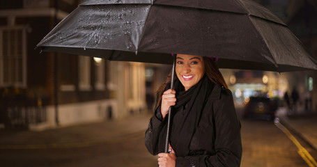 Pretty Latina woman standing under umbrella on rainy night