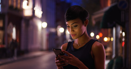 Pretty smiling African American female reads text message on the street at night