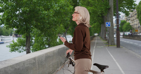 Active senior Caucasian woman on bike looking at river and texting on phone