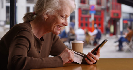 Happy senior woman shopping online with credit card and phone in coffee shop