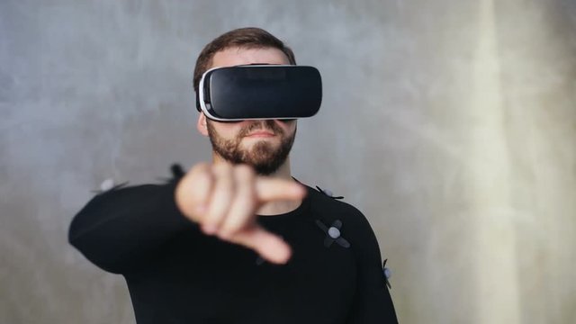 indoors portrait of attractive adult man in motion-capture suit with motion sensors and virtual reality glasses holding hand up and swapping excited experience of using hitech wearable device studio
