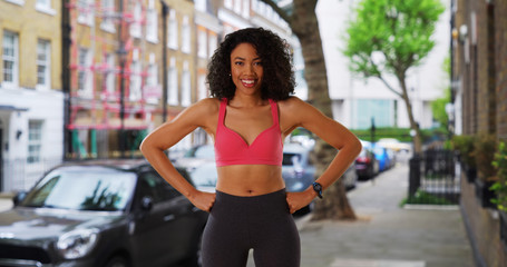 Fototapeta na wymiar Cheerful smiling black female jogger in active wear standing on city street