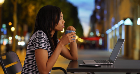 Pretty black woman on city street at night drinking coffee and using laptop