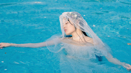 Beautiful fairy in a beautiful white wedding dress and garlands of flowers, standing in swimming pool with sunny ripples on turquoise water, beautiful Chinese girl in water in sunny day. 