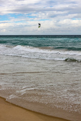 Obraz premium Tide rolling onto the shore with horizon under cloud shadow and kite surfer out in the water - vertical
