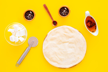 Raw rolled out pizza dough near cheese mozzarella, olive oil, spices, knife for pizza on yellow background top view mockup