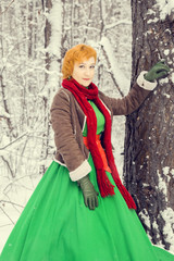 fiery red-haired woman in a ball green dress with a red leather belt in the costume of dwarf assistant Santa Claus in the winter forest with huge candy, a chest of gifts, the concept of the new year
