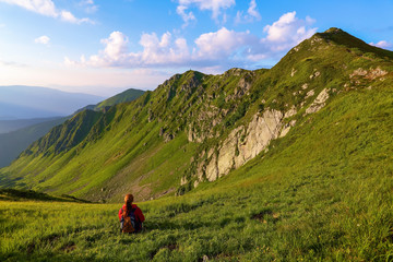 Fototapeta na wymiar The view with the high rocky mountains. A young girl sits on the green grass among the flowers. Sur rays. Beautiful clouds are slowly moving in the sky. Summer scenery.