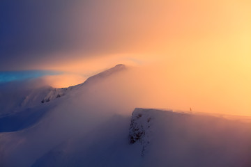 At the edge of the snowy rock someone is standing. The high mountains in the fog, morning sky is enlighten with the orange colored light in winter day.