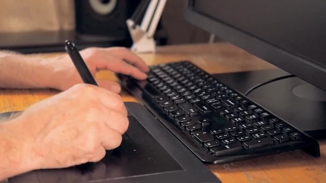 Close up view on graphic designer's hands using tablet and touch screen pen for drawing. One hand clicking buttons on keyboard, another one drawing with pen. Blurred background, soft selective focus