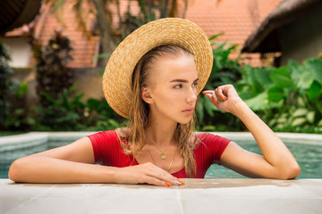 Close up portrait of a beautiful young woman in sexy red swimsuit and vintage straw hat in pool posing to camera. Hot sunny day. Tropic island vacation. Summer travel girl, active hipster lifestyle.