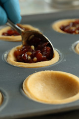 Person putting mince pie filling onto pastry base in a metal baking tray with a spoon
