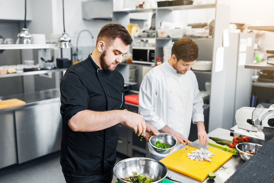 cooking food, profession and people concept - happy male chef and cook with knife chopping champignons and adding greens to salad bowl at restaurant kitchen