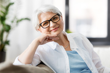 vision, age and people concept - portrait of happy senior woman in glasses sittin on sofa at home