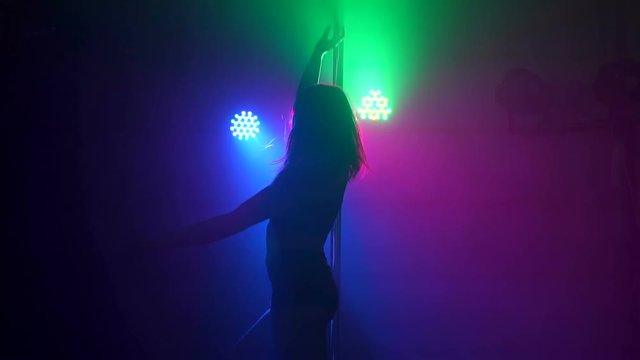 Gorgeous flexible girl dances a sensual dance on a pylon in the dark with multicolored searchlights and smoke.