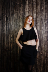 Fototapeta premium Portrait of a beautiful redheaded girl in black top and black skirt posing in the studio next to the wooden wall.