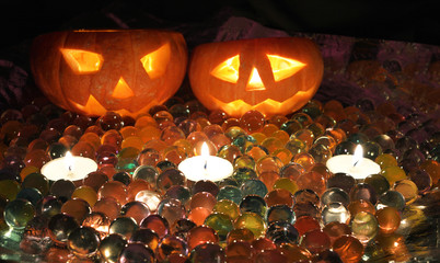 Two orange glowing carved pumpkin heads for the holiday of Helloon, surrounded by colorful jellied balls, with three candles, lie on a table on a dark background.