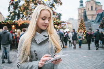 Fototapeta premium A beautiful young blonde woman or tourist uses a tablet next to a Christmas tree during the Christmas holidays at the Old Town Square in Prague, Czech Republic.
