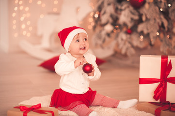 Obraz na płótnie Canvas Funny baby girl wearing santa claus suit playing with christmas decor in room over lights. Winter season.
